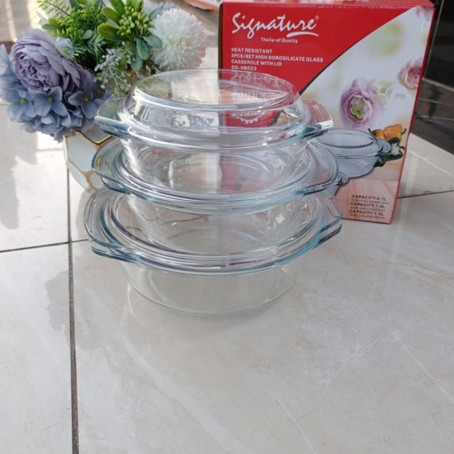A stack of three clear borosilicate glass casserole dishes with lids, displayed on a kitchen counter with a product box in the background.