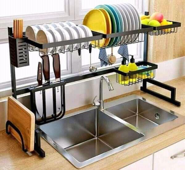 An over-the-sink dish drainer in silver color, mounted above a kitchen sink, holding plates, cups, and kitchen utensils.