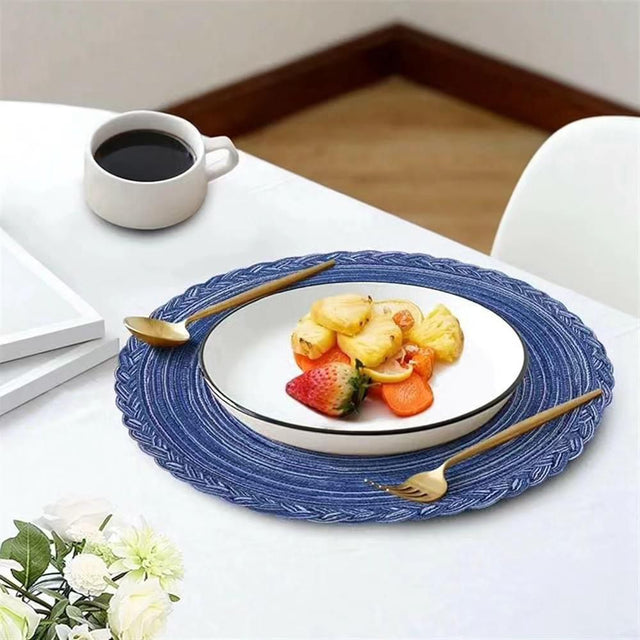 A blue braided round table mat is placed on a white surface, with a white plate of fruit and a cup of coffee adjacent to it.