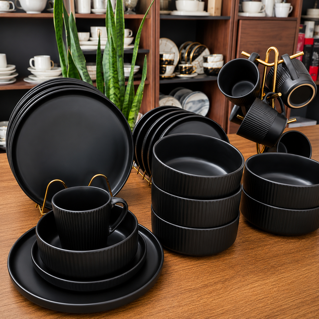 Elegant matte black dinnerware set with plates, bowls, mugs, and gold-tone mug stand displayed on a wooden table in a home décor showroom