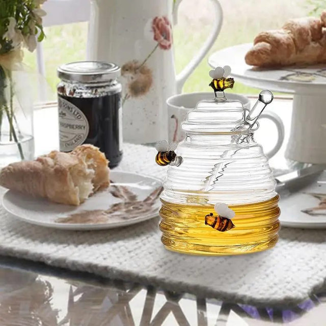 A clear glass honey dispenser with a honeycomb pattern and a dipper, placed on a table with food and drinks in the background.
