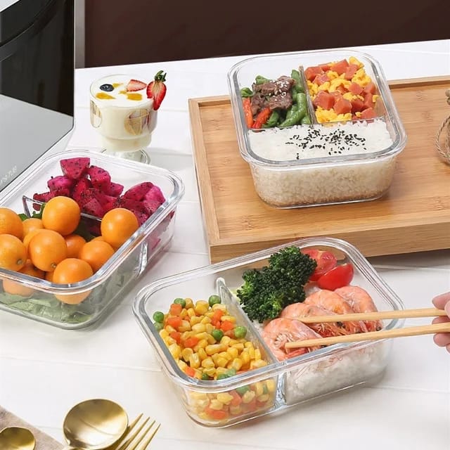 Partitioned glass lunch boxes with food compartments, one with blue lids and one with clear silicone lids, on a kitchen counter.