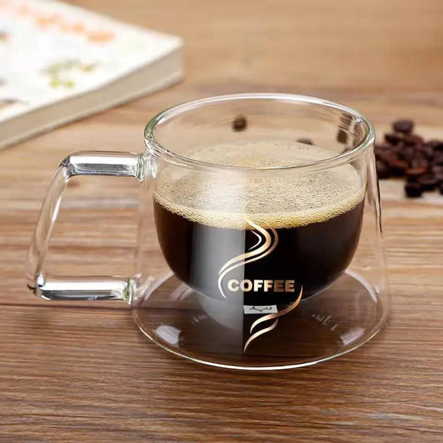 A double-walled clear glass espresso cup with 'COFFEE' text on it, filled with coffee, placed on a wooden surface with coffee beans scattered around.