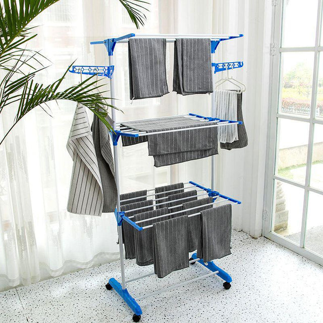 A three-layer portable cloth drying rack with blue frames and silver pipes, equipped with wheels and containing some folded grey towels, set against a home backdrop with a plant.
