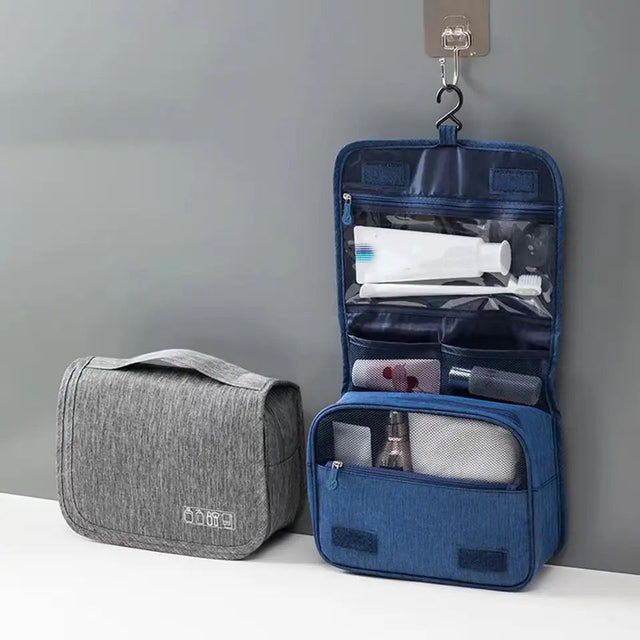 A foldable cosmetic bag in gray color, displayed open with various toiletries like a toothbrush, razor, and bottles arranged inside it, hanging from a hook on the wall.
