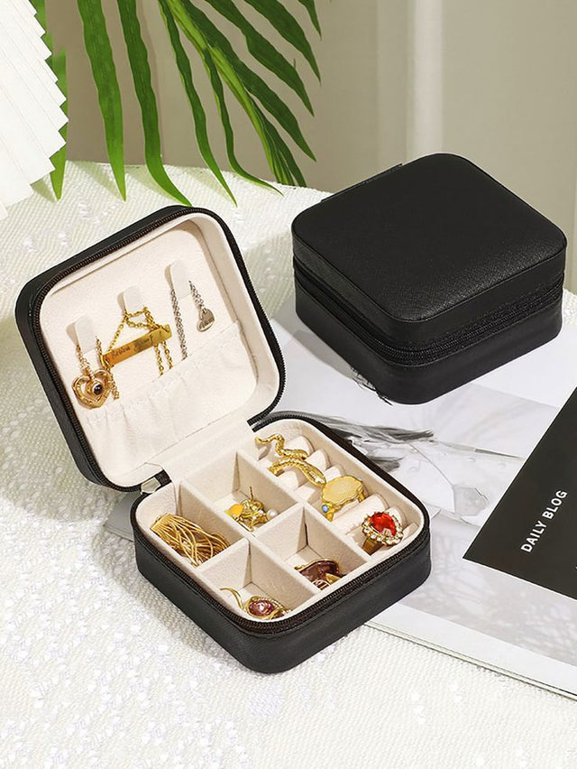 Black jewelry box with compartments containing gold jewelry on a white surface with greenery in the background.