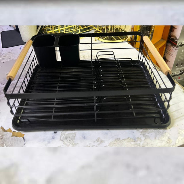 A black metal dish rack with wooden handle and multiple compartments, including a section for utensils, placed on a kitchen countertop.