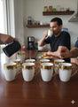 People enjoying coffee in a kitchen with multiple white mugs with gold accents on a table.
