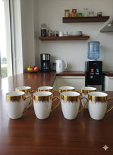 Set of white mugs with gold trim on a wooden table in a kitchen.
