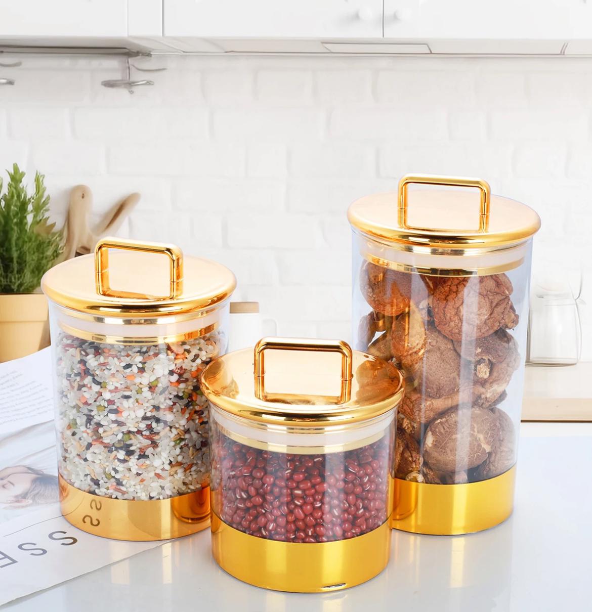 Three clear acrylic storage jars with gold-colored lids, containing different dry food items, placed on a kitchen counter.