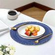 A blue braided round table mat is placed on a white surface, with a white plate of fruit and a cup of coffee adjacent to it.
