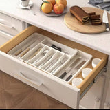 A white expandable cutlery drawer organizer with various compartments, placed inside an open kitchen drawer.