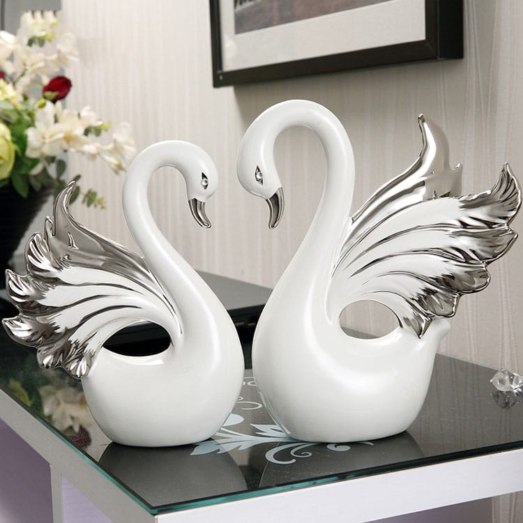 A pair of Nordic ceramic swan ornaments with white bodies and silver wings, placed on a glass surface.