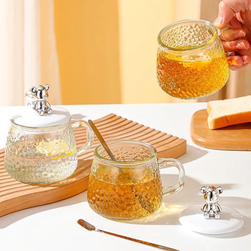 A set of two clear glass cups with one cup being filled with a yellow beverage and the other cup having a silver lid, placed on a wooden board.