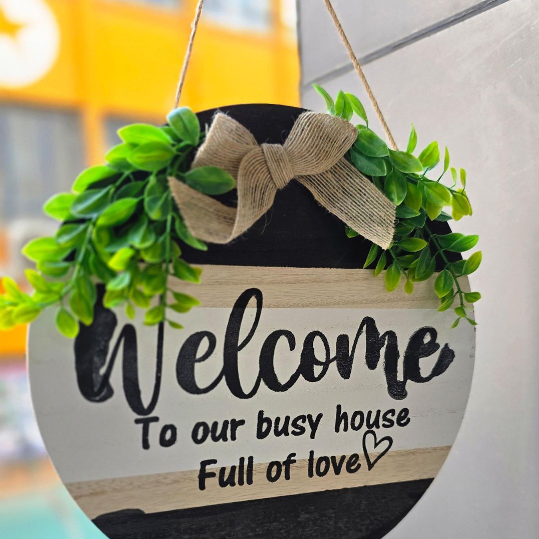 A wooden sign with a black and white background, green leaves, and a bow, with the text 'Welcome to our busy house full of love'.