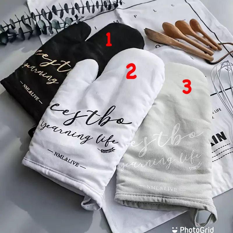 Three pairs of oven gloves, two black and one white, with text on the cuffs, displayed on a table with a pot holder and wooden utensils in the background.
