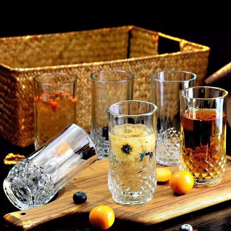A set of three glass lowball glasses with a diamond pattern, filled with different colored beverages, placed on a wooden board next to a wicker basket.