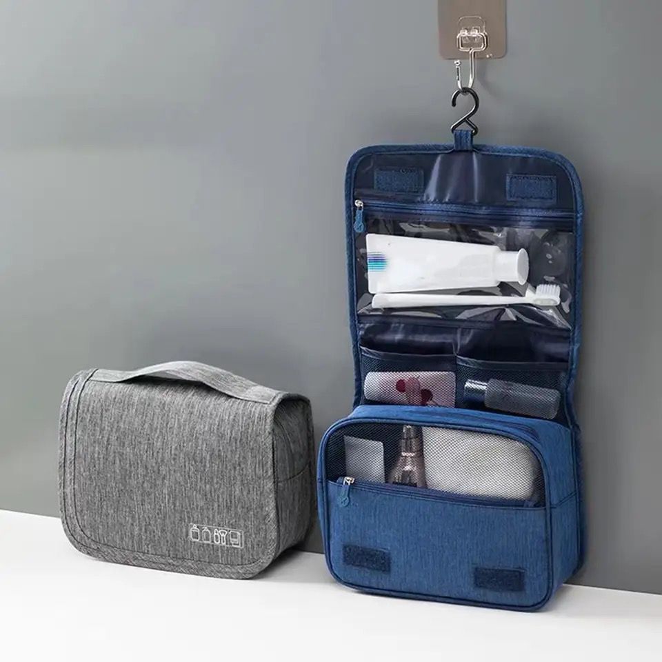 A foldable cosmetic bag in gray color, displayed open with various toiletries like a toothbrush, razor, and bottles arranged inside it, hanging from a hook on the wall.