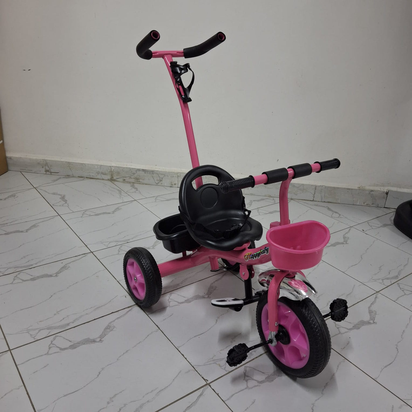 A pink tricycle with a black seat and wheels, featuring a push handle on a white tiled floor.