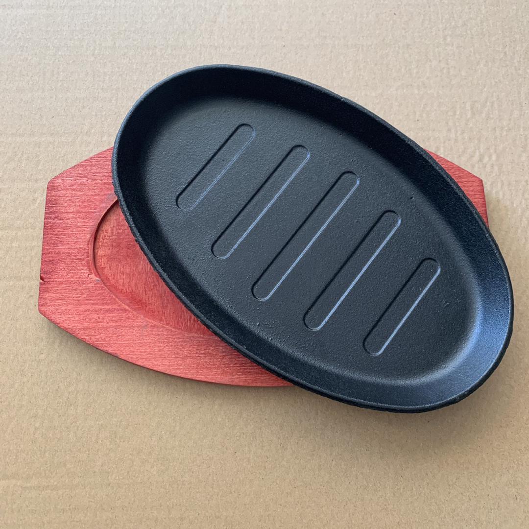 A black sizzling plate with grooves, accompanied by a red base, likely made of cast iron, set against a beige background.