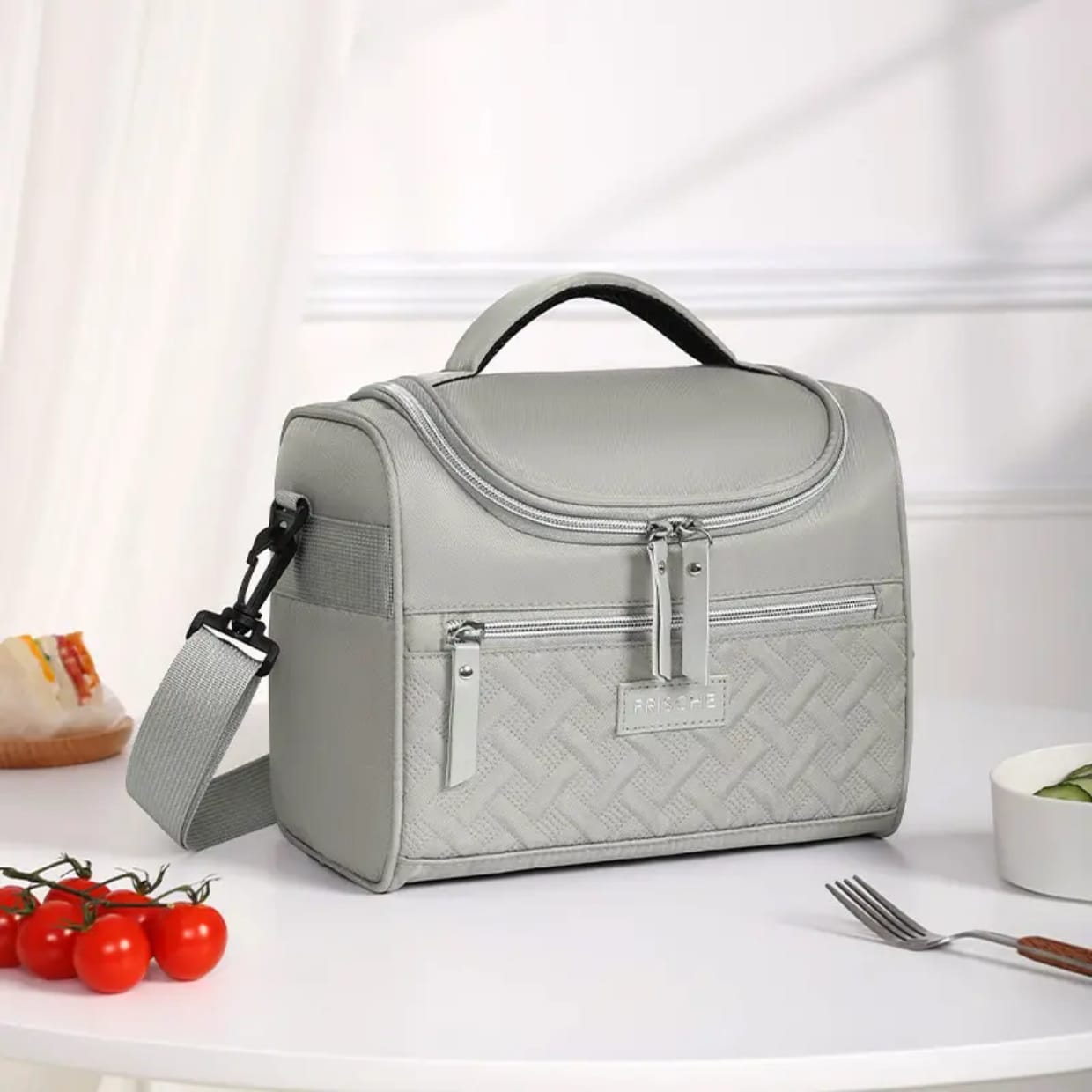 A gray insulated lunch bag with a quilted design, featuring a front zipper pocket and a side handle, placed on a table with food items around it.