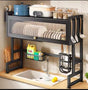 Black steel over-sink kitchen rack with dust-proof dish cabinet, top shelf for appliances, side utensil hooks, and mid-level wire basket mounted above a kitchen sink